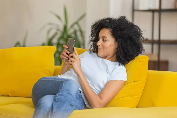 Retrato de una joven afroamericana enviando mensajes de texto o viendo información en su teléfono. Morena con el pelo rizado sentado en un sofá amarillo en una habitación luminosa en casa. De cerca.. — Foto de Stock