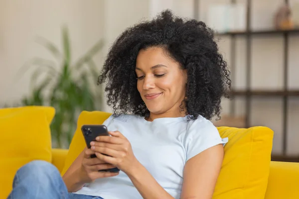 Retrato de una joven afroamericana enviando mensajes de texto o viendo información en su teléfono. Morena con el pelo rizado sentado en un sofá amarillo en una habitación luminosa en casa. De cerca.. — Foto de Stock