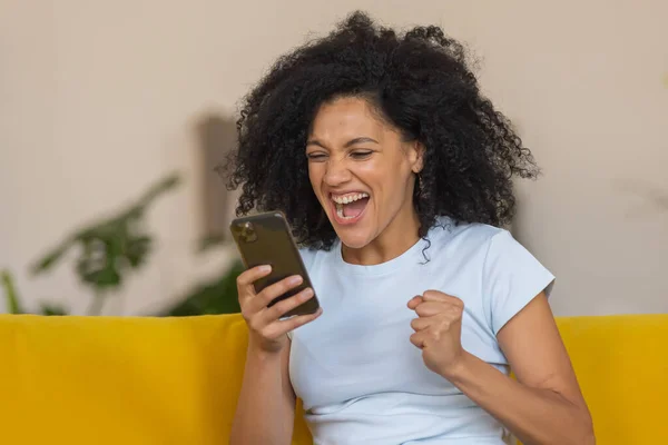 Retrato de una joven afroamericana mirando un teléfono inteligente se regocija con entusiasmo por las buenas noticias o la información recibida. Morena sentada en un sofá amarillo en una habitación luminosa. De cerca.. — Foto de Stock