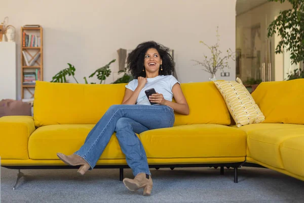 スマートフォンを見ている若いアフリカ系アメリカ人女性の肖像画は、受信した良いニュースや情報で熱狂的に喜びます。ブルネットともに巻き毛の上に座っている黄色のソファで明るい家の部屋 — ストック写真