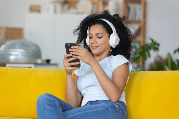 Retrato de una joven afroamericana que disfruta de la música con grandes auriculares blancos usando un teléfono inteligente. Morena con el pelo rizado sentado en un sofá amarillo en una habitación luminosa en casa. De cerca.. —  Fotos de Stock