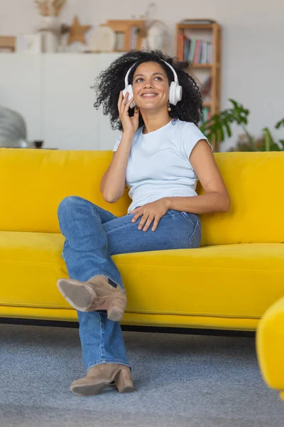 一个年轻的非洲裔美国女人的肖像喜欢大的白色耳机的音乐。留着卷曲头发的布鲁内特坐在一间明亮的房间里的黄色沙发上. — 图库照片