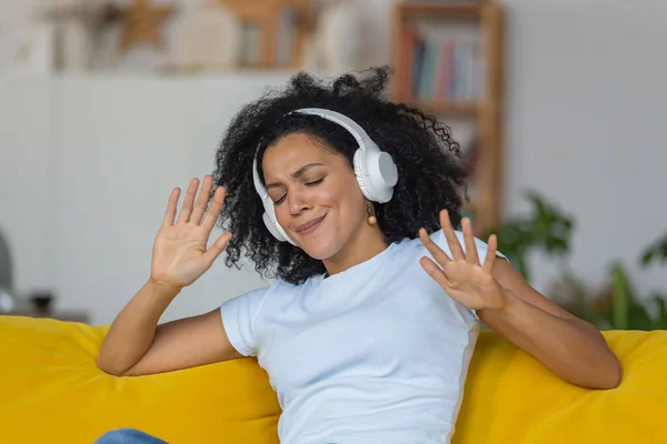 Porträtt av en ung afroamerikansk kvinna njuter av musik med stora vita hörlurar. Brunett med lockigt hår sittande på gul soffa i ett ljust hemrum. Närbild. — Stockfoto