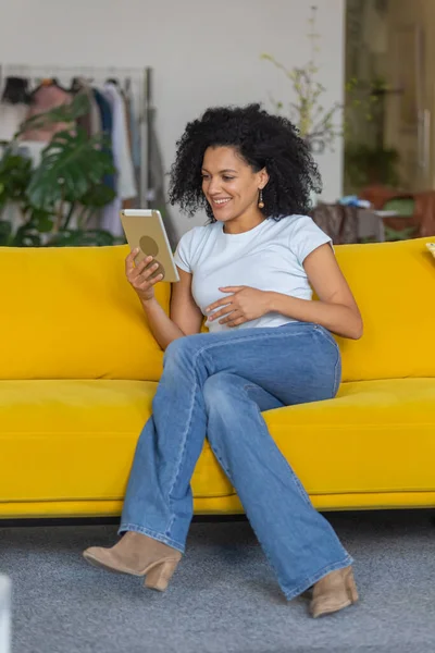 Retrato de uma jovem afro-americana falando em uma videochamada em seu tablet digital. Morena com cabelo encaracolado sentado no sofá amarelo em uma sala de casa brilhante. — Fotografia de Stock