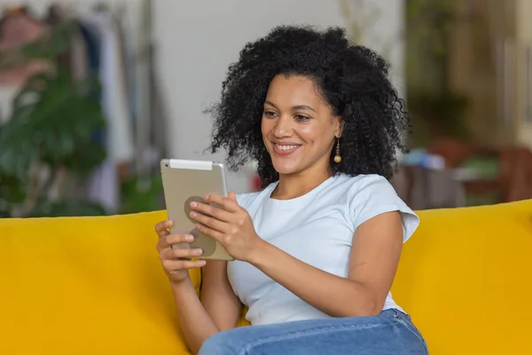 Retrato de una joven afroamericana hablando en una videollamada en su tableta digital. Morena con el pelo rizado sentado en un sofá amarillo en una habitación luminosa en casa. De cerca.. — Foto de Stock