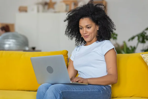 Portret młodej Afroamerykanki piszącej na klawiaturze laptopa. Brunetka z kręconymi włosami siedząca na żółtej kanapie w jasnym salonie. Zamknij się.. — Zdjęcie stockowe