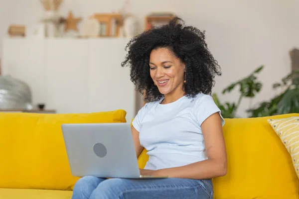 Retrato de uma jovem afro-americana falando em uma videochamada no laptop portátil. Morena com cabelo encaracolado sentado no sofá amarelo em uma sala de casa brilhante. Fechar. — Fotografia de Stock