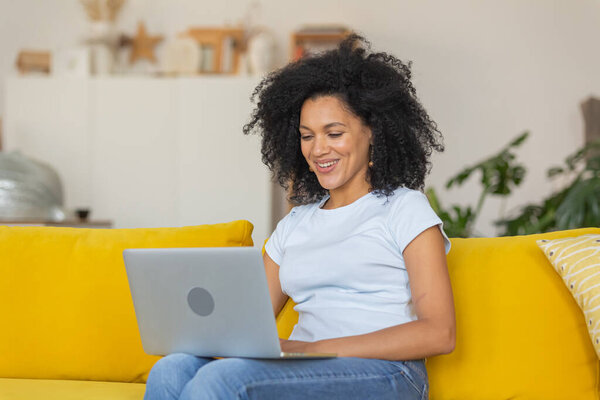 Портрет молодой афроамериканки, разговаривающей на видео-вызове на портативном ноутбуке. Брюнетка с вьющимися волосами сидит на жёлтом диване в яркой домашней комнате. Закрыть.