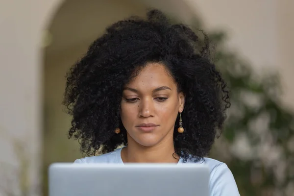 Ritratto di una giovane donna afroamericana che digita sulla tastiera di un computer portatile. Bruna con capelli ricci seduta su un divano giallo in una luminosa stanza di casa. Da vicino.. — Foto Stock