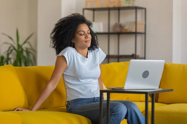 Retrato de uma jovem afro-americana cansada sentada com os olhos fechados perto do laptop. Morena com cabelos cacheados posando no sofá amarelo em uma sala de casa brilhante. Fechar. — Fotografia de Stock