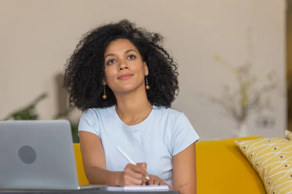 Genç bir Afrikalı Amerikalı kadının portresi video konferansında dizüstü bilgisayar kullanarak konuşur ve not defterine notlar alır. Sarı koltukta oturan kıvırcık saçlı esmer, parlak bir ev odasında oturuyor. Kapat.. — Stok fotoğraf