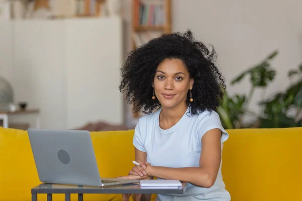 Ritratto di una giovane donna afroamericana parla in videoconferenza con un computer portatile e prende appunti in un quaderno. Bruna con capelli ricci seduta su un divano giallo in una luminosa stanza di casa. Da vicino.. — Foto Stock