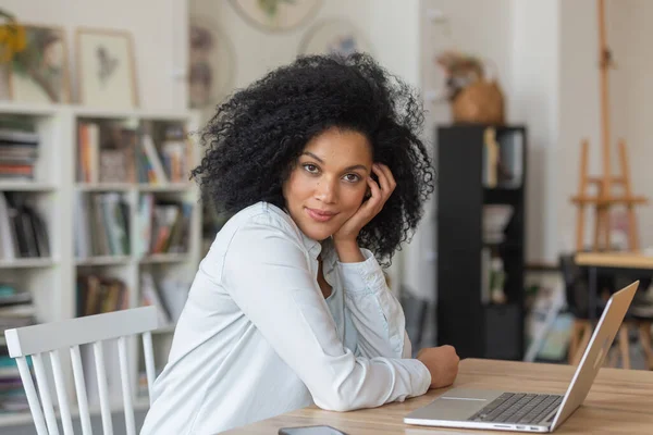 Portrait de jeune femme afro-américaine regardant la caméra et souriant. Brunette aux cheveux bouclés en chemisier blanc s'assoit à table dans un bureau à domicile léger. Enseignement à distance ou concept de travail à distance. Gros plan. — Photo