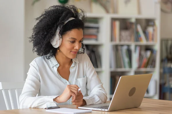 Portrait d'une femme afro-américaine parlant par vidéoconférence à l'aide d'un ordinateur portable et d'écouteurs prenant des notes sur un bloc-notes. Brunette est assise à table au bureau à la maison. Gros plan. — Photo