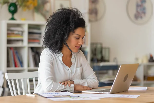 Portrait d'une jeune femme afro-américaine parlant par vidéoconférence à l'aide d'un ordinateur portable et d'un casque. Brunette aux cheveux bouclés en chemisier blanc s'assoit à table dans un bureau à domicile léger. Gros plan. — Photo