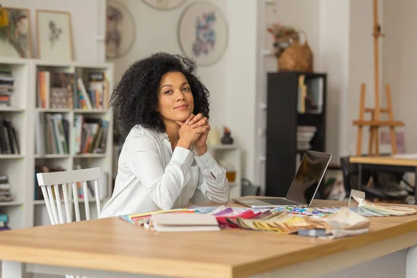 Ritratto di una giovane donna afroamericana che guarda la macchina fotografica. Il designer femminile in camicetta bianca si siede a un tavolo in ufficio vicino a tavolozza a colori e campioni di tessuti per un progetto di progetto. Da vicino.. — Foto Stock