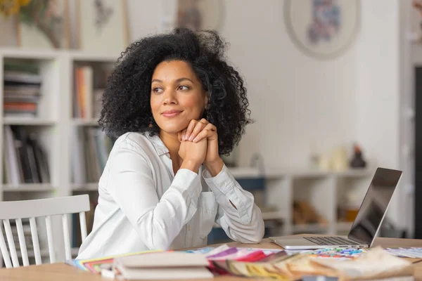 Portrait d'une jeune afro-américaine regardant vers le côté. Femme designer en chemisier blanc s'assoit à une table au bureau près de la palette de couleurs et des échantillons de tissus pour un projet de design. Gros plan. — Photo