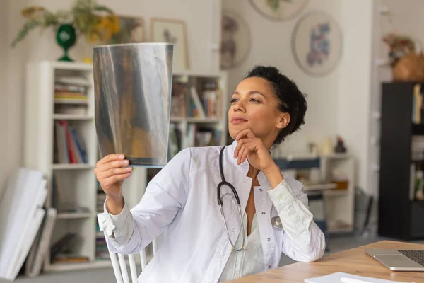 En ung kvinnlig läkare undersöker en ögonblicksbild av lungorna. Afro amerikanska kvinna sitter vid bordet på ett sjukhus kontor. Telemedicin. Läkarbesök online. Närbild. — Stockfoto