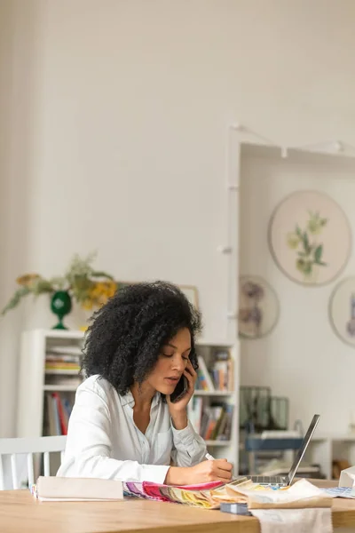 非洲裔美国女青年的画像，通过彩色调色板和面料样品，通过电话讨论项目的设计。女设计师坐在灯光办公室的桌子旁。靠近点. — 图库照片