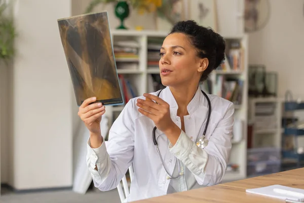 En ung kvinnlig läkare undersöker en ögonblicksbild av lungorna. Afro amerikanska kvinna sitter vid bordet på ett sjukhus kontor. Telemedicin. Läkarbesök online. Närbild. — Stockfoto