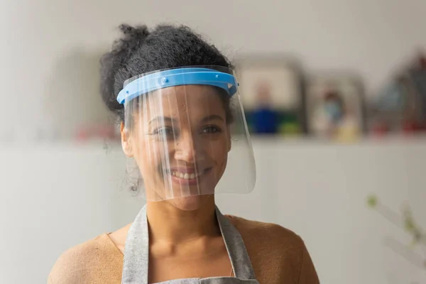 Mulher afro-americana bonita olhando para o lado usando uma máscara plástica protetora e sorrindo. Raça mista jovem posando contra o pano de fundo de uma sala de luz. Fechar. — Fotografia de Stock