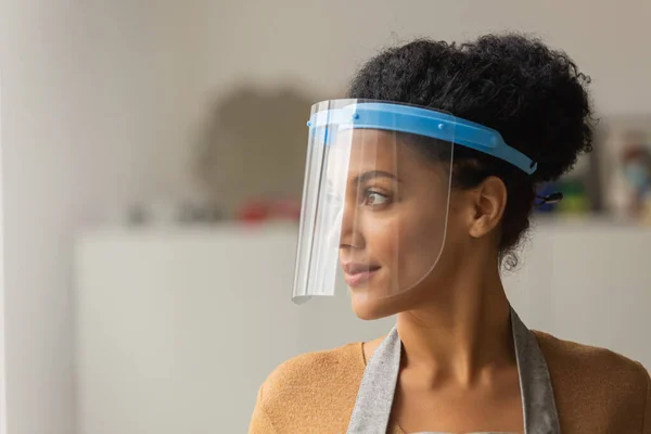 Mujer afroamericana bonita mirando hacia un lado con una máscara protectora de plástico y sonriendo. Mujer joven de raza mixta posando sobre el telón de fondo de una sala de luz. De cerca.. — Foto de Stock