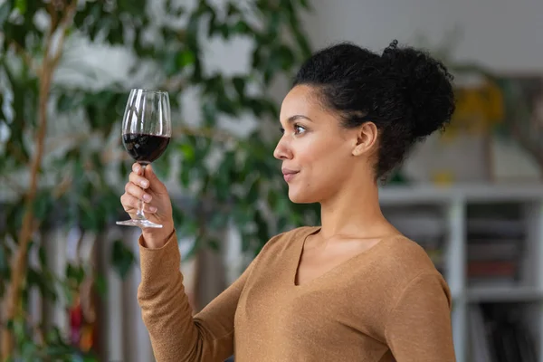 Portrait of a young African American woman looking at a glass of red wine. Cute mixed race female posing against blurred background of light room with green plants. Close up. — Stock Photo, Image