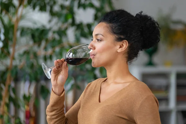 Porträtt av en ung afroamerikansk kvinna som dricker rött vin från glasbägare. Söt blandras hona njuter av smak av dryck mot suddig bakgrund av ljus rum med gröna växter. Närbild. — Stockfoto