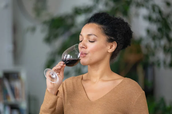 Retrato de uma jovem afro-americana bebendo vinho tinto de cálice de vidro. Fofa mista fêmea está gostando de gosto de bebida contra fundo borrado de sala de luz com plantas verdes. Fechar. — Fotografia de Stock