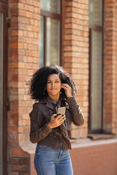 携帯電話を手にカメラを見てスタイリッシュな若いアフリカ系アメリカ人女性の肖像画。黒レンガ造りの建物を背景に路上でポーズ茶色の革のジャケットのブルネット. — ストック写真