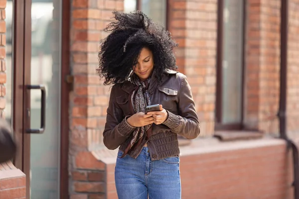 Retrato de una joven afroamericana enviando mensajes de texto en su smartphone. Morena con pelo rizado en chaqueta de cuero y jeans elegantes posando en la calle contra el telón de fondo del edificio de ladrillo borroso. — Foto de Stock