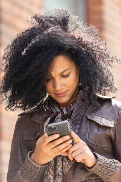 Retrato de una joven afroamericana enviando mensajes de texto en su smartphone. Morena con el pelo rizado en chaqueta de cuero y vaqueros elegantes posando en la calle contra el telón de fondo del edificio de ladrillo borroso. De cerca.. — Foto de Stock