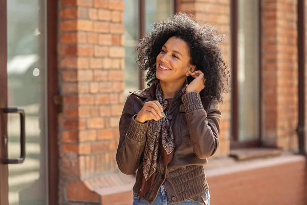 若いアフリカ系アメリカ人女性の肖像画かわいい笑顔。黒レンガ造りの建物の背景を背景に路上でポーズを革のジャケットに巻き毛のブルネット。閉じろ!. — ストック写真
