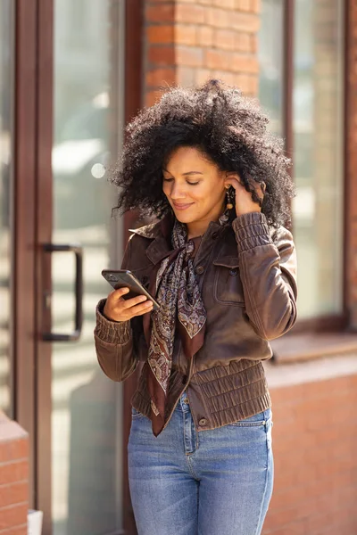 スマートフォンにアフリカ系アメリカ人の若い女性の肖像画と笑顔。黒レンガ造りの建物を背景に、革のジャケットに巻き毛のブルネットとストリートにポーズをとるスタイリッシュなジーンズ — ストック写真