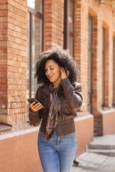 スマートフォンにアフリカ系アメリカ人の若い女性の肖像画と笑顔。黒レンガ造りの建物を背景に、革のジャケットに巻き毛のブルネットとストリートにポーズをとるスタイリッシュなジーンズ — ストック写真