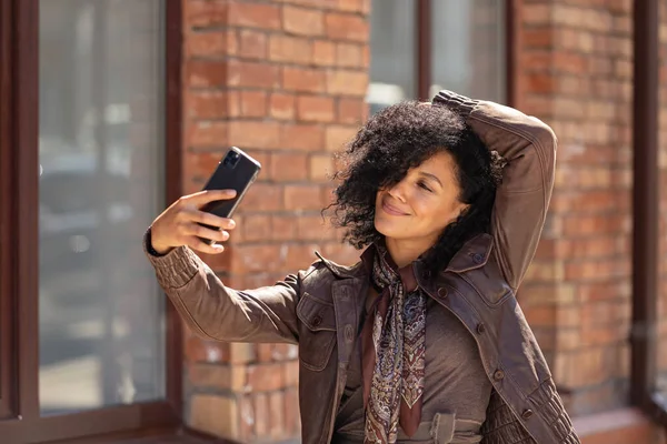 Retrato de la joven mujer afroamericana con estilo haciendo selfie en el teléfono móvil. Morena con el pelo rizado en chaqueta de cuero marrón posando en la calle contra el telón de fondo del edificio de ladrillo borroso. De cerca.. — Foto de Stock