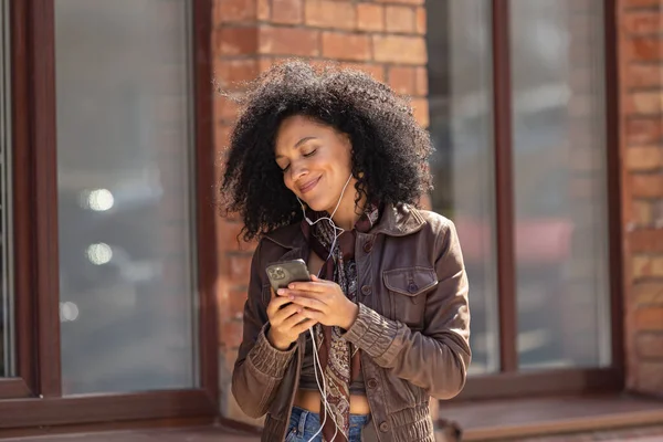 若いアフリカ系アメリカ人女性のポートレートは、携帯電話を使用してヘッドフォンで音楽を楽しんでいます。黒レンガ造りの建物の背景を背景に路上でポーズを革のジャケットに巻き毛のブルネット。閉じろ!. — ストック写真