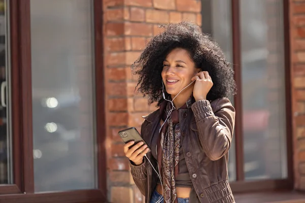 若いアフリカ系アメリカ人女性のポートレートは、携帯電話を使用してヘッドフォンで音楽を楽しんでいます。黒レンガ造りの建物の背景を背景に路上でポーズを革のジャケットに巻き毛のブルネット。閉じろ!. — ストック写真