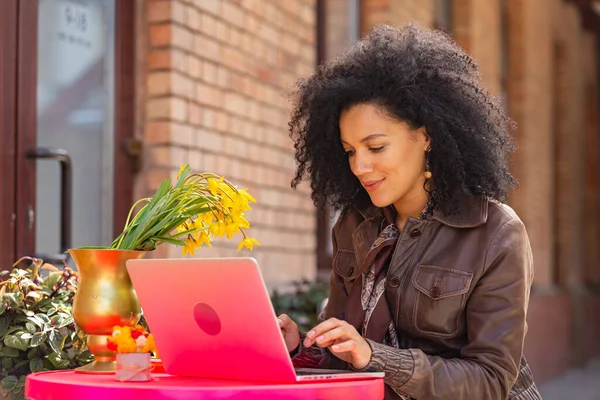 Portrait de la jeune femme afro-américaine élégante tapant sur un clavier d'ordinateur portable. Brune veste en cuir marron assise à une table dans un café confortable dans la rue. Gros plan. — Photo