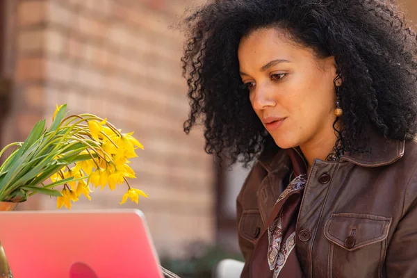 Portrait de la jeune femme afro-américaine élégante tapant sur un clavier d'ordinateur portable. Brune veste en cuir marron assise à une table dans un café confortable dans la rue. Gros plan. — Photo