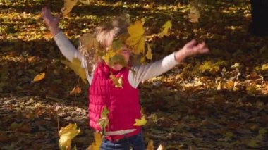 Pembe yelekli mutlu küçük bir kız gülüyor ve bir kucak dolusu altın dökülen yaprakları fırlatıyor. Güneşli bir günde sonbahar ormanında eğlence. Aile hafta sonu dışarıda. Ağır çekim. Kapat..