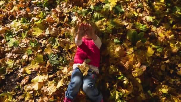 A menina jaz no chão espalhada com folhas verdes e amarelas caídas. A criança cobre os olhos com a palma da mão da luz solar brilhante. Movimento lento. Fechar. — Vídeo de Stock