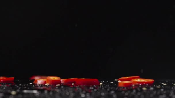Plátky červené horké chilli papričky padající na mokrý povrch na černém pozadí studia. Kousky kořeněné zeleniny, přísada na hořké jídlo. Veganské tapety na jídlo do restaurace. Zavřít. Zpomalený pohyb. — Stock video