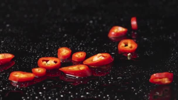 Rodajas de chile rojo picante cayendo sobre una superficie húmeda de color negro. Trozos de verduras picantes, un ingrediente para el plato amargo. Papel pintado de comida vegana para restaurante. De cerca. Movimiento lento. — Vídeo de stock