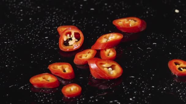 Rodajas de chile rojo picante cayendo sobre una superficie húmeda de color negro. Trozos de verduras picantes, un ingrediente para el plato amargo. Papel pintado de comida vegana para restaurante. De cerca. Movimiento lento. — Vídeo de stock