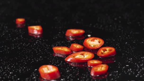 Rodajas de chile rojo picante cayendo sobre una superficie húmeda de color negro. Trozos de verduras picantes, un ingrediente para el plato amargo. Papel pintado de comida vegana para restaurante. De cerca. Movimiento lento. — Vídeos de Stock