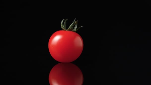 Un pomodoro rosso maturo con una coda verde ruota su una superficie del tavolo da cucina nera riflettente al rallentatore. Secco lucido vegetale estivo filatura da vicino su uno sfondo nero studio. — Video Stock