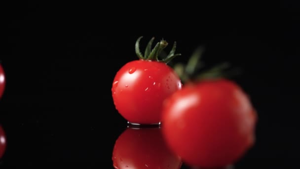 Tomates rojos maduros con una cola verde en gotas de agua rotan sobre una superficie reflectante negra de la mesa de la cocina en cámara lenta. Verduras húmedas y brillantes de verano girando de cerca sobre un fondo de estudio negro. — Vídeo de stock