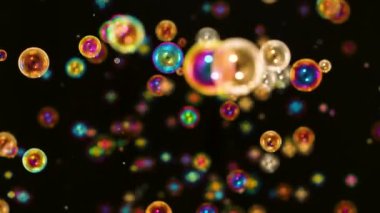 Birçok gökkuşağı parlak sabun köpükleri siyah arka planda uçar ve patlar. Çok renkli yuvarlak kabarcıklar uzayda ağır çekimde hareket eder ve ışık ışınlarında parıldar. Kapat..