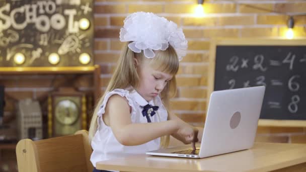 Klein meisje zit aan tafel en typt op het laptop toetsenbord op de achtergrond van een schoolklas. Een basisschoolmeisje met witte strikken in de les. School, thuisonderwijs, afstandsonderwijs. — Stockvideo